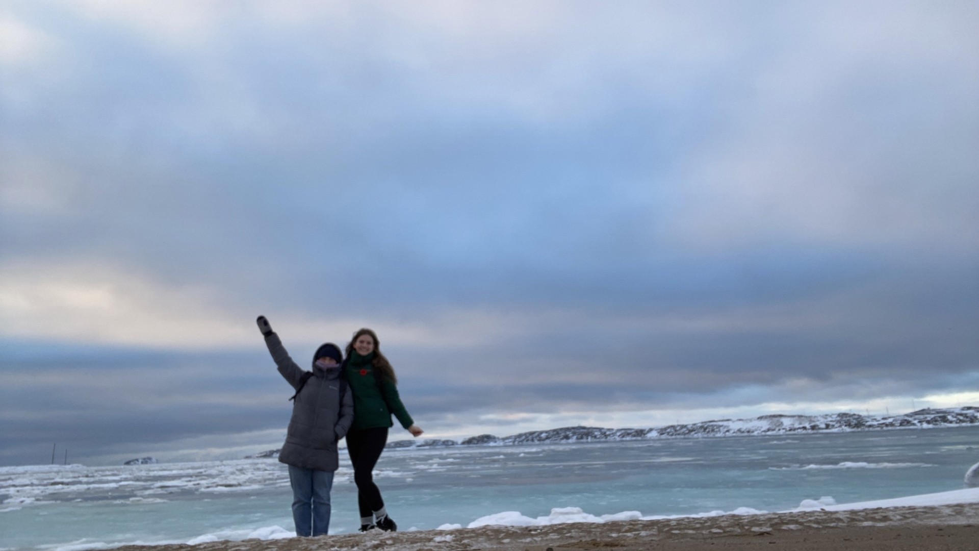 Two people in jackets standing on a beach waving