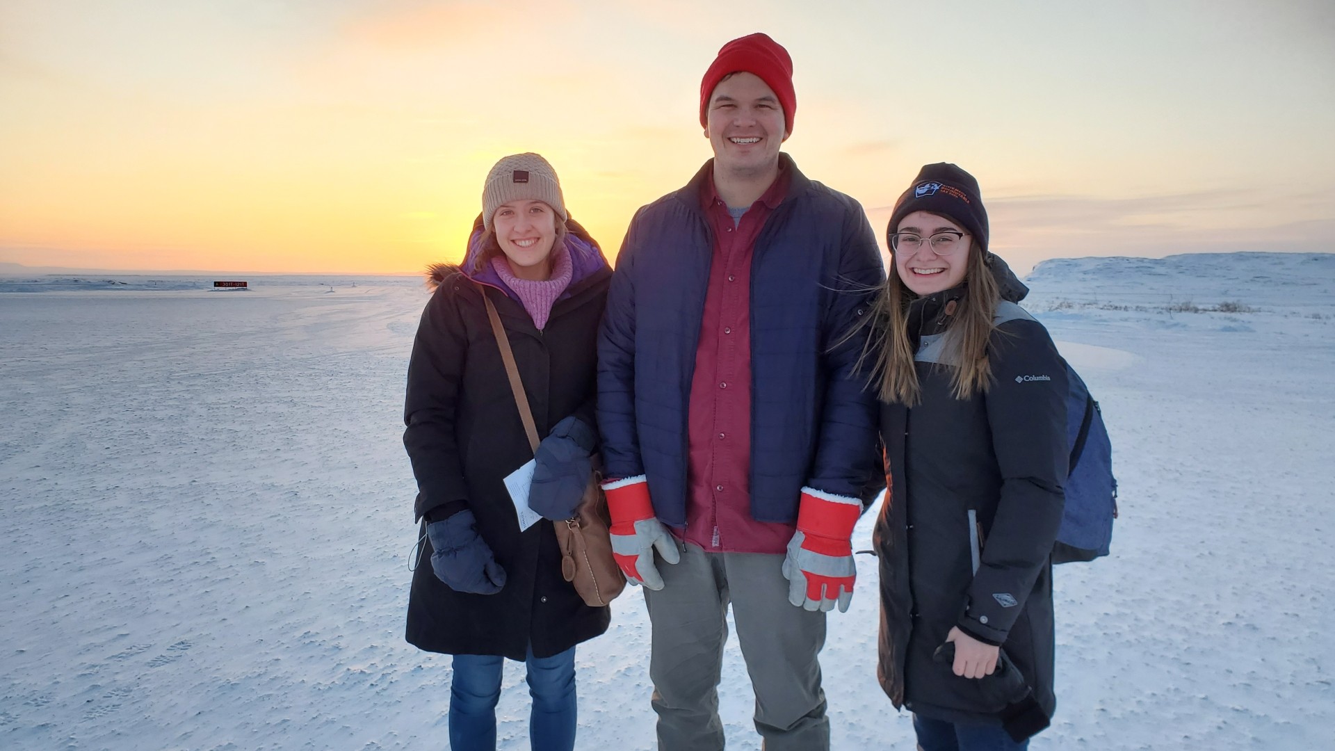 Three people standing outside on snowy ground with a sunrise or sunset in the background