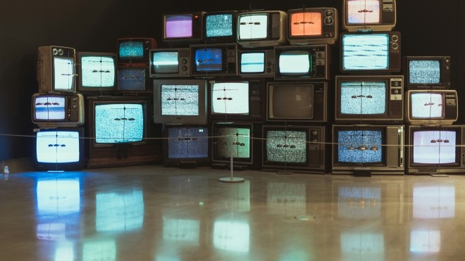 Bank of multiple old box TVs piled on top of each other. Their screens are different colours. The screens' reflect off the shiny beige floor.