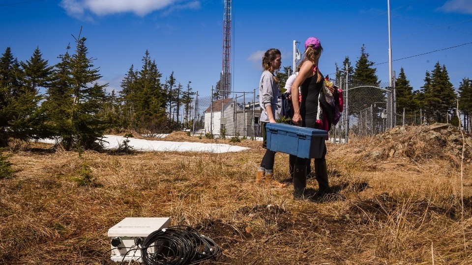 Two individuals carrying a container in a field with equipment around.