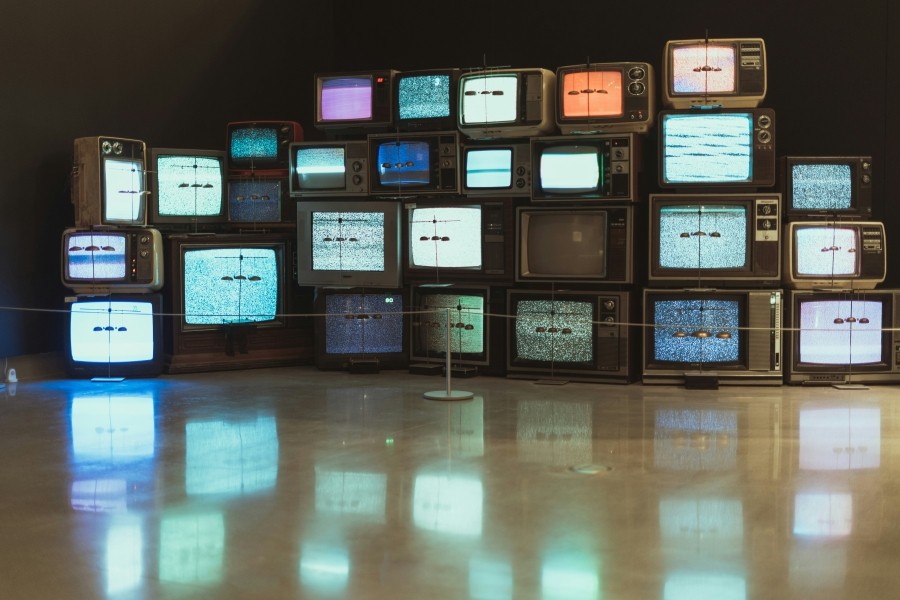 Bank of multiple old box TVs piled on top of each other. Their screens are different colours. The screens' reflect off the shiny beige floor.