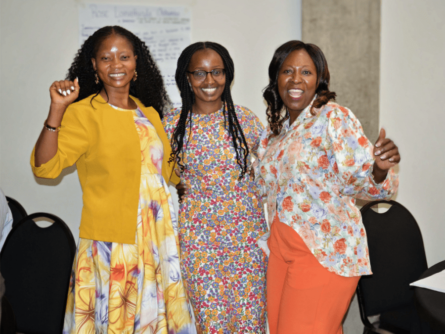 Three smiling Black women pose arm-in-arm indoors, wearing colorful dresses and tops; their raised hands and joyful expressions convey celebration.