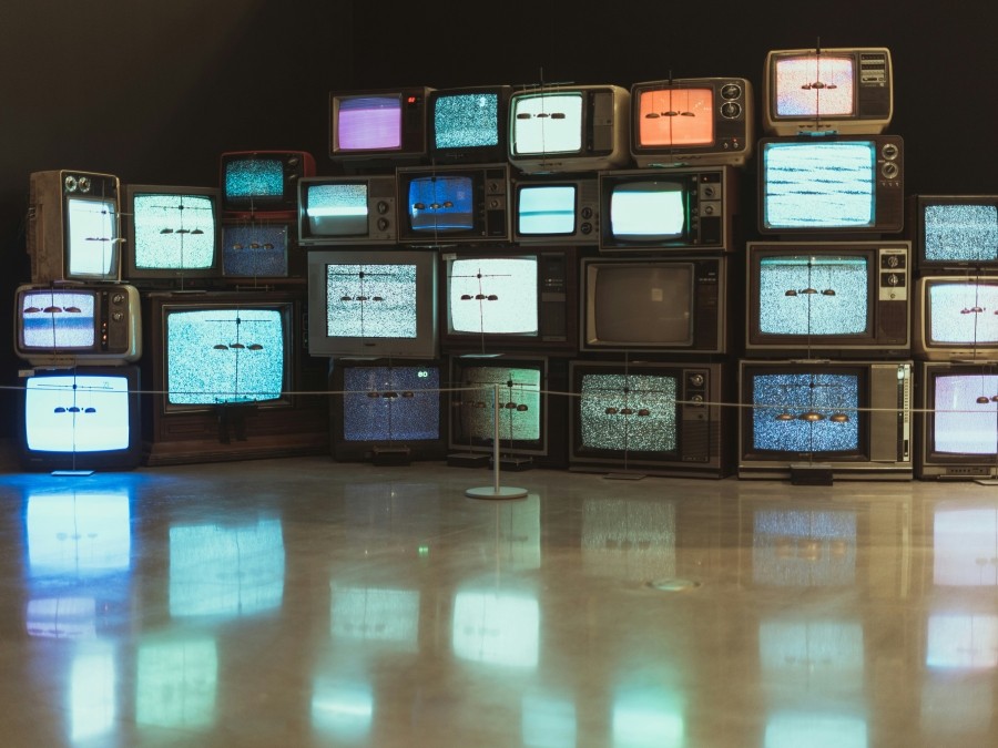 Bank of multiple old box TVs piled on top of each other. Their screens are different colours. The screens' reflect off the shiny beige floor.